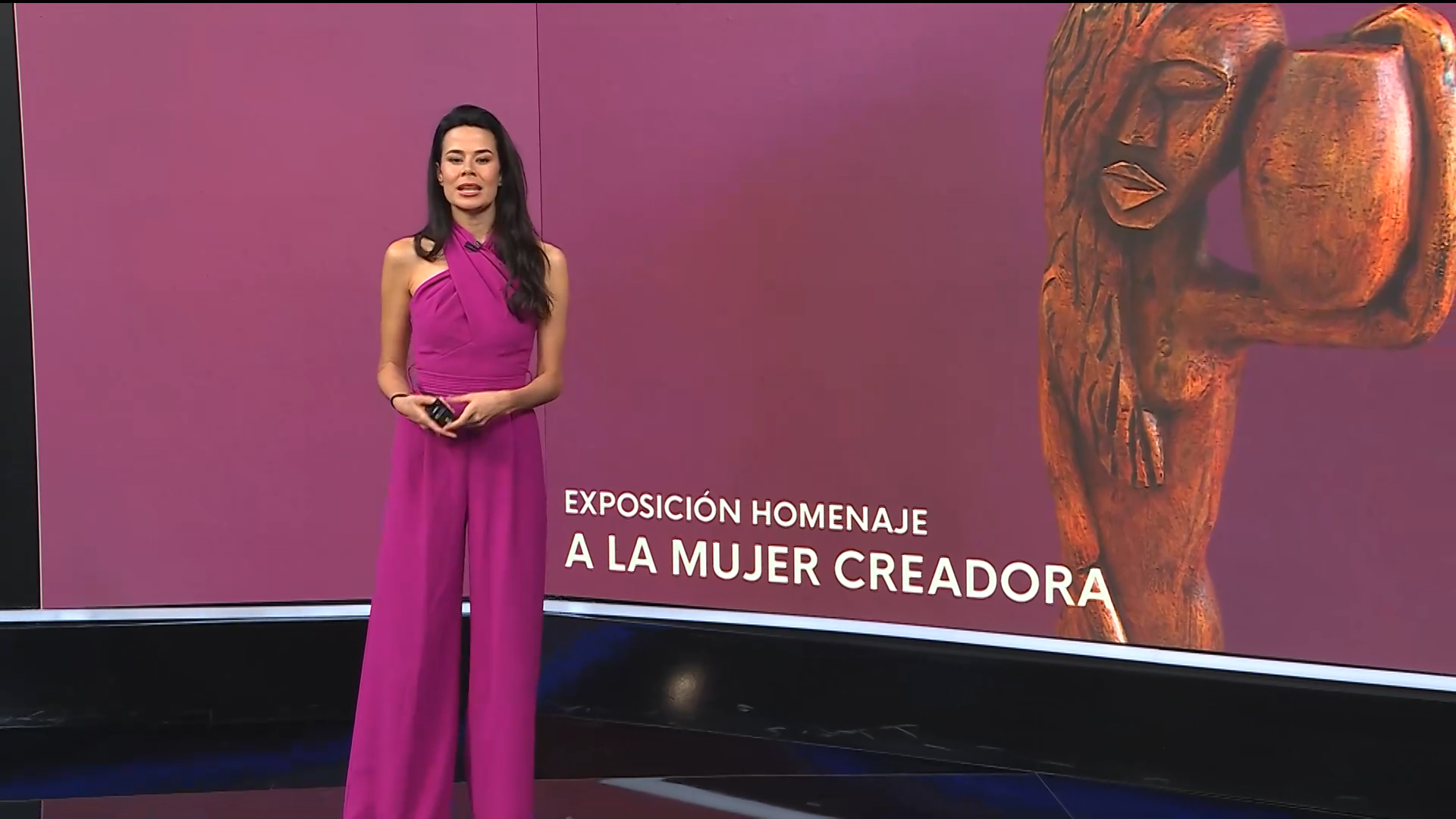 Captura de vídeo de la emisión de un programa: una presentadora, de pie, frente a una pantalla con una escultura.