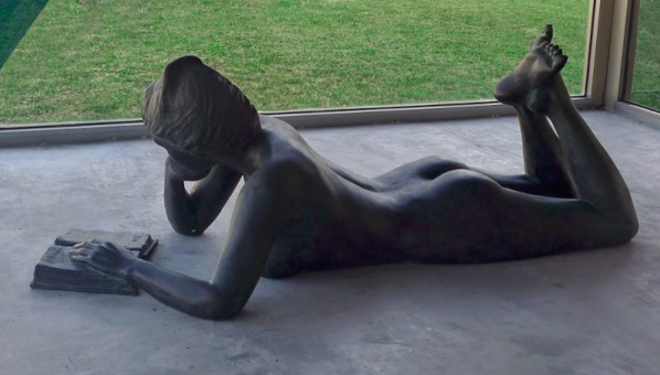 Estatua de mujer desnuda acostada boca abajo leyendo un libro, con una mano en la cara.