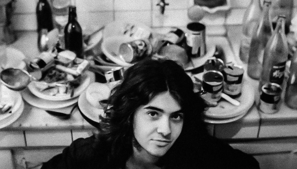 Fotografía de adolescente con pelo largo frente a mesa de cocina desbordada de restos.