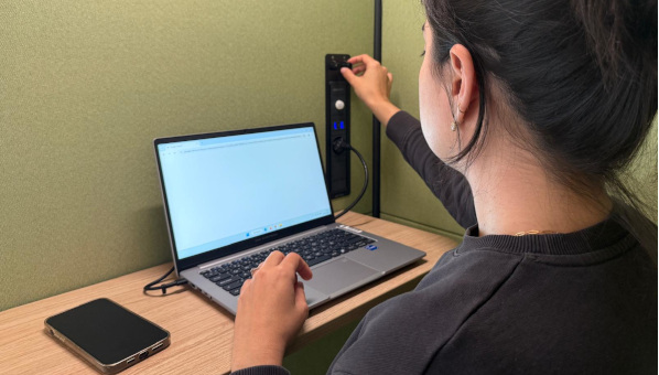 Mujer sentada en cabina frente a un pequeño estante, con ordenador portátil y móvil, maneja un mando en la pared de la cabina