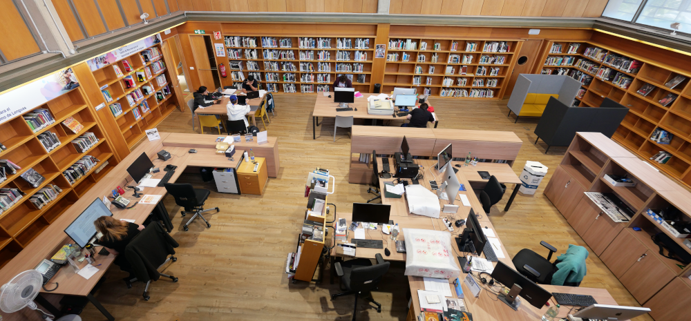 Sala de biblioteca con estantes de libros en las paredes y gente estudiando o trabajando en mesas.