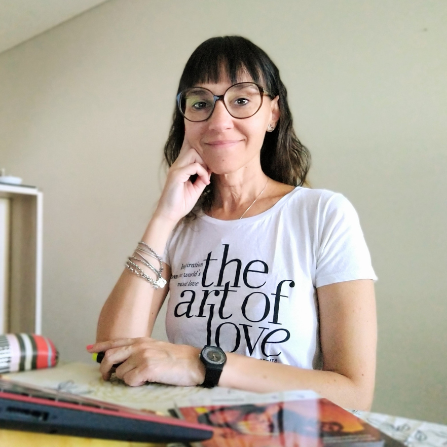 Mujer con gafas posa sonriente sentada frente a mesa de trabajo con útiles. Su camiseta pone el mensaje The art of love.