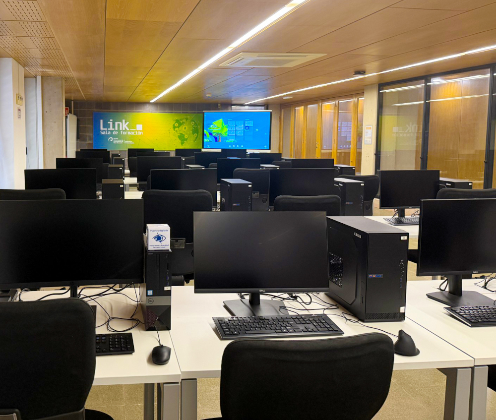Sala de formación con puestos de ordenadores en varias filas, y con pizarra digital.