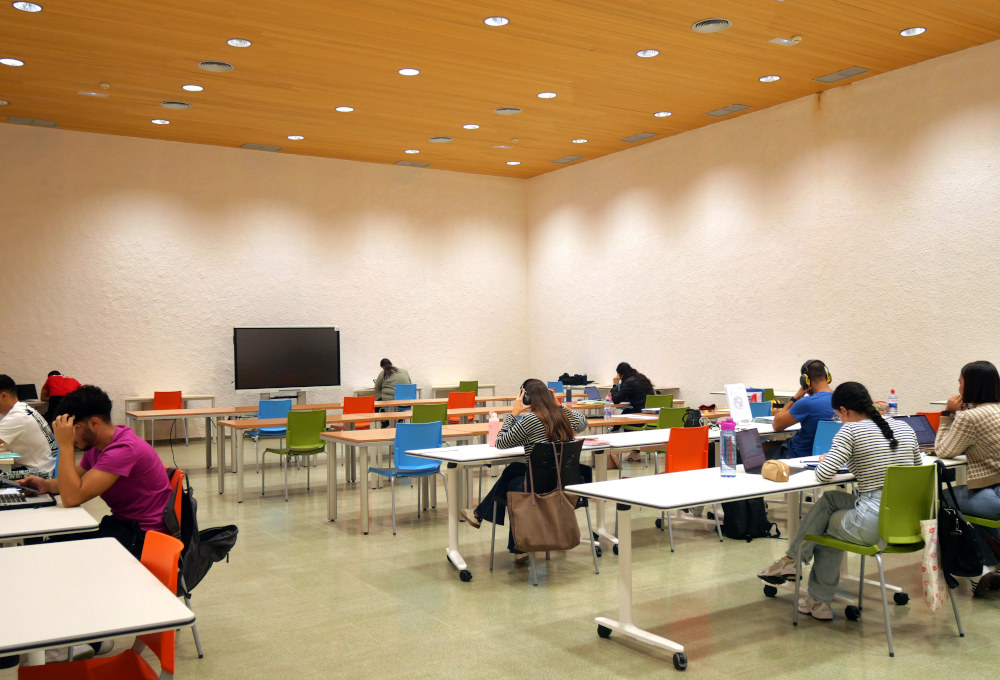 Vista de sala amplia con estudiantes en mesas blancas y sillas de colores variados.