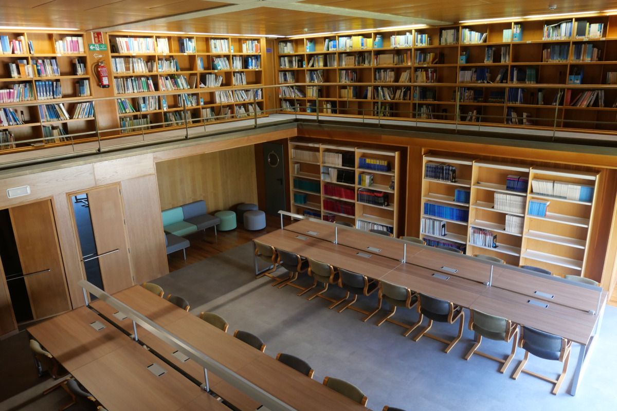 Vista de sala de biblioteca con dos plantas de estanterías. En la planta baja, dos filas de mesas equipadas. En la planta alta, corredor junto a pared con estantes.