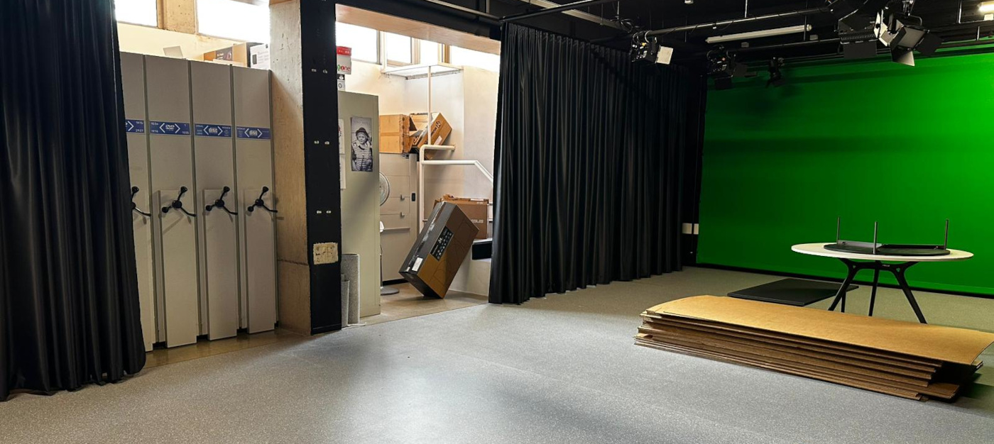 Vista de sala con estanterías compactas, escenario con croma verde, mesa, cortinas negras y focos en el techo.