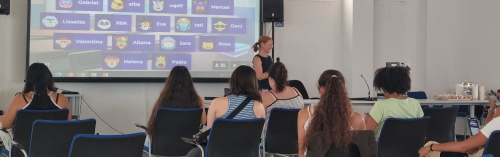 Grupo de chicas estudiantes sentadas en aula atienden a profesora con micro. En pantalla se proyecta una actividad didáctica.