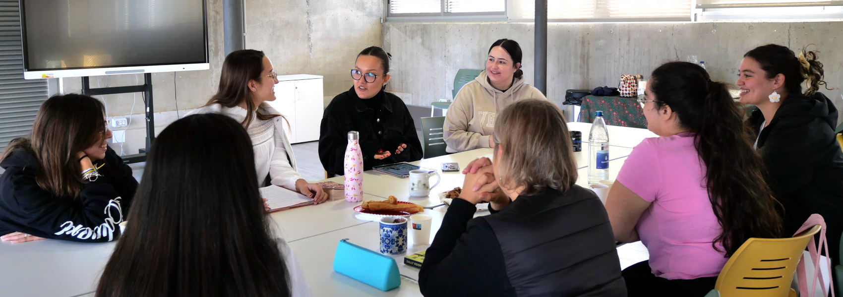 Varias mujeres jóvenes conversan en torno a una mesa con libros, bebidas y comida.