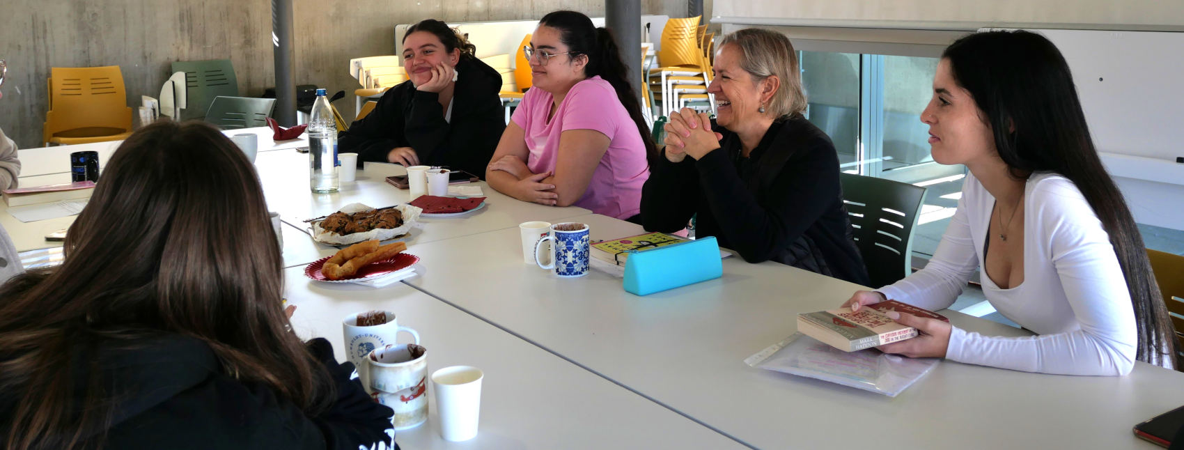 Vista de varias mujeres sentadas ante una mesa conversando con libros, comidas y bebidas.