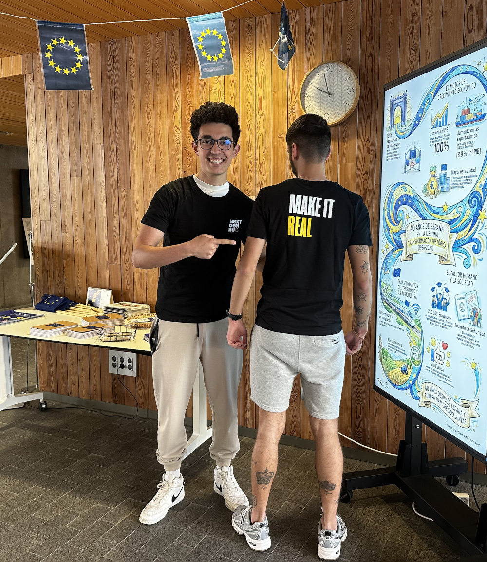 Dos estudiantes con camisetas promocionales de la Unión Europea posan; uno sonríe a la cámara mientras apunta con el dedo a otro, de espaldas, mostrando el texto de la camiseta «MAKE IT REAL».