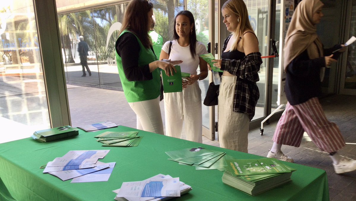 Dos estudiantes reciben información de una voluntaria de la campaña frente a una mesa con folletos, a la entrada de un edificio