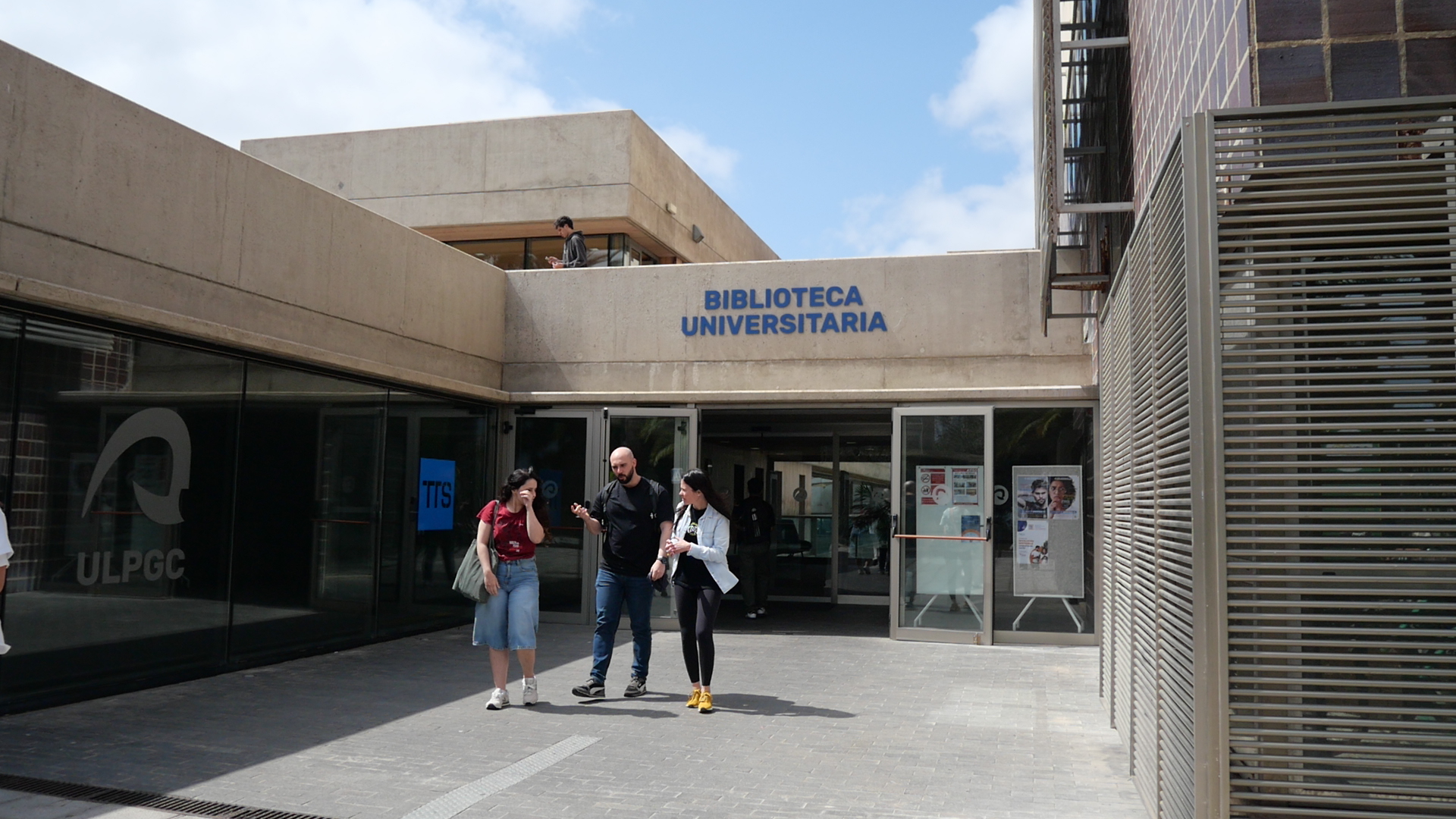 Tres personas saliendo de edificio con rótulo Biblioteca Universitaria