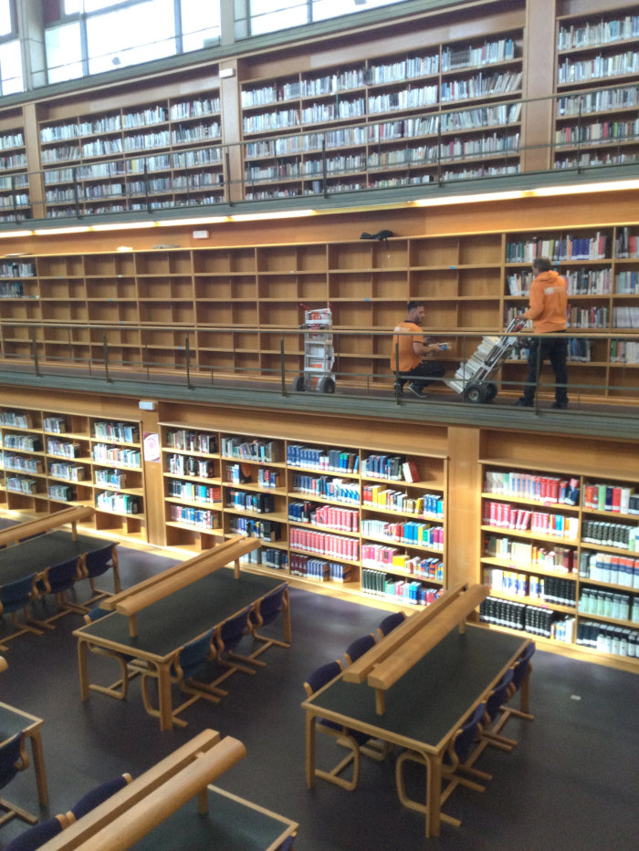 Vista de sala de biblioteca con 3 plantas de estantes de libros donde dos operarios con carretilla vacían estantes.