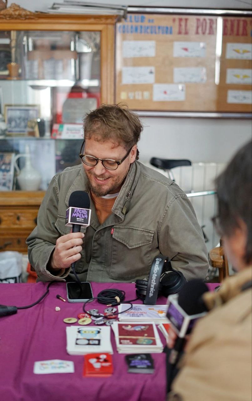 Un hombre sonriente locuta con un micrófono en mano sentado ante una mesa frente a otra persona con micrófono en mano