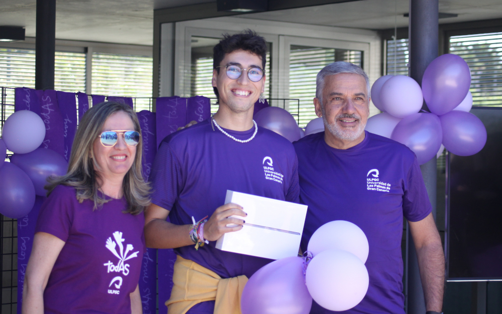 Tres personas: directora de la Unidad de Igualdad, persona ganadora sosteniendo el premio -tableta-, y rector, todas posando con camiseta violeta.