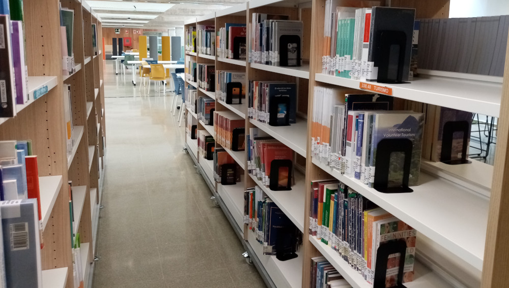 Vista de pasillo en perspectiva entre estantes de libros de una biblioteca, con mesas y sillas al fondo.