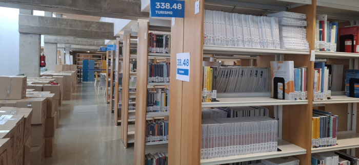 Estantes de libros en biblioteca con el rótulo "338.48 Turismo". Cajas acumuladas en pasillo.