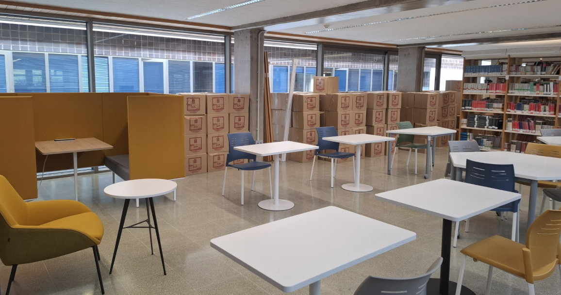 Vista de sala de biblioteca con mesas de estudio, sillas, estantes de libros y cajas acumuladas.