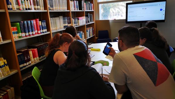 Vista de estudiantes en torno a una mesa de trabajo, con una gran pantalla, entre estantes de una biblioteca.