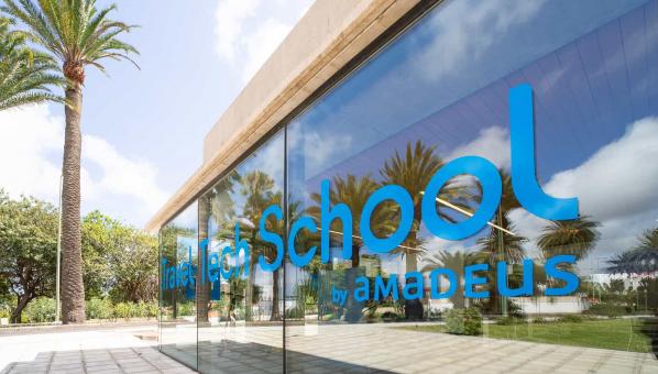 Talleres Travel Tech School ULPGC by Amadeus Vista del exterior de la Travel Tech School: una pared completamente acristalada con el logotipo de la TTS que refleja el exterior del Edificio Central de la Biblioteca Universitaria, donde destacan muchas palmeras. Al fondo, una palmera y más árboles.