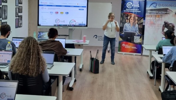 Una profesora habla a unas 8 personas sentadas en mesas de a dos, con ordenador, en una sala docente.