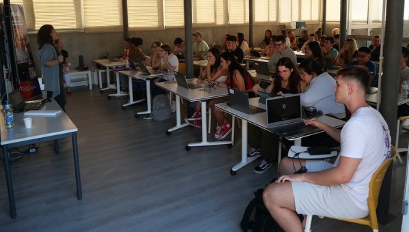 Más de treinta estudiantes atienden explicaciones de una profesora sentadas ante mesas mientras consultan sus ordenadores.