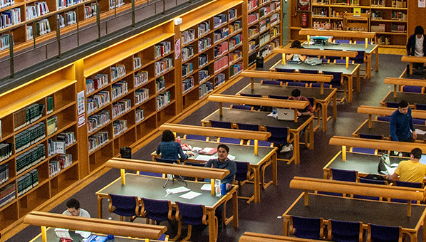 Vista parcial de sala de biblioteca con libros en estantes de varios pisos y personas estudiando en mesas alineadas en dos filas.