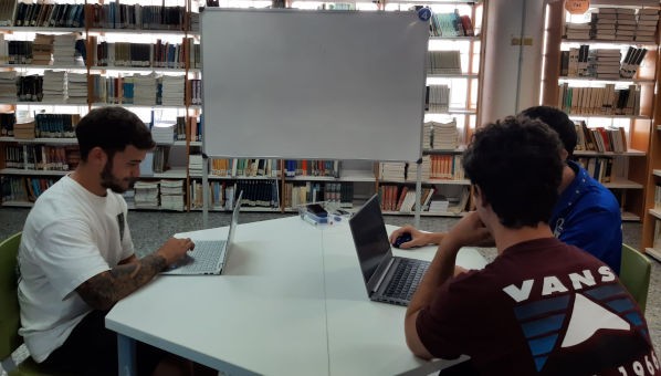 Tres alumnos estudian con ordenadores portátiles en una mesa de una biblioteca.