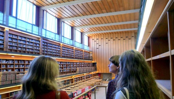 Tres personas visitan una sala principal de lectura de biblioteca con tres pisos de estantes.