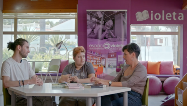 Tres personas dialogan sentadas ante una mesa en una biblioteca.