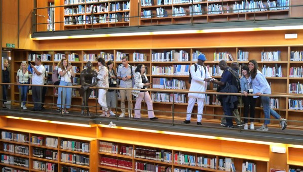 Un grupo de personas visita una biblioteca desde el segundo nivel de una sala de lectura de 3 niveles de estantes de libros.