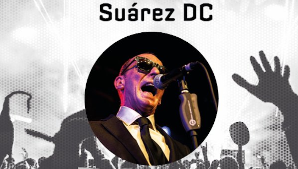 Hombre cantando ante un micro con gafas de sol, chaqueta y corbata. De fondo: siluenta de personas asistentes a un concierto.