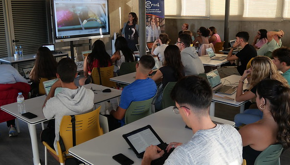 Aula con estudiantes ante mesas con ordenadores y tabletas personales, atendiendo la explicación de una profesora que se apoya en una pizarra digital.