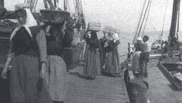Fotografía antigua de mujeres desestibando, con cestas sobre la cabeza, pesca de barcos veleros en un muelle.