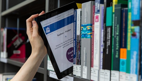 Una mano de mujer entresaca de un estante de una biblioteca un libro electrónico