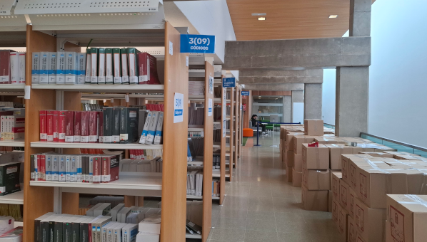 Vista de estantes de libros en una sala con cajas acumuladas. En un cartel indicativo se lee "3(09) Códigos"