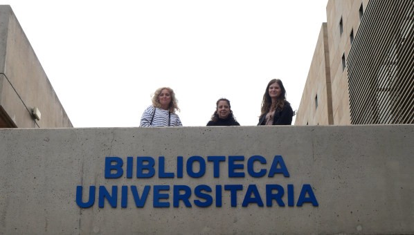 Tres mujeres posan asomadas a la terraza de un edificio de hormigón sobre el rótulo Biblioeca Universitaria