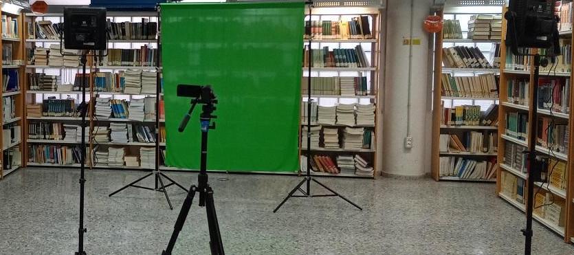Espacio de grabación Vista de una cámara de vídeo y dos focos en trípodes frente a una tela verde para croma, entre estantes de una biblioteca