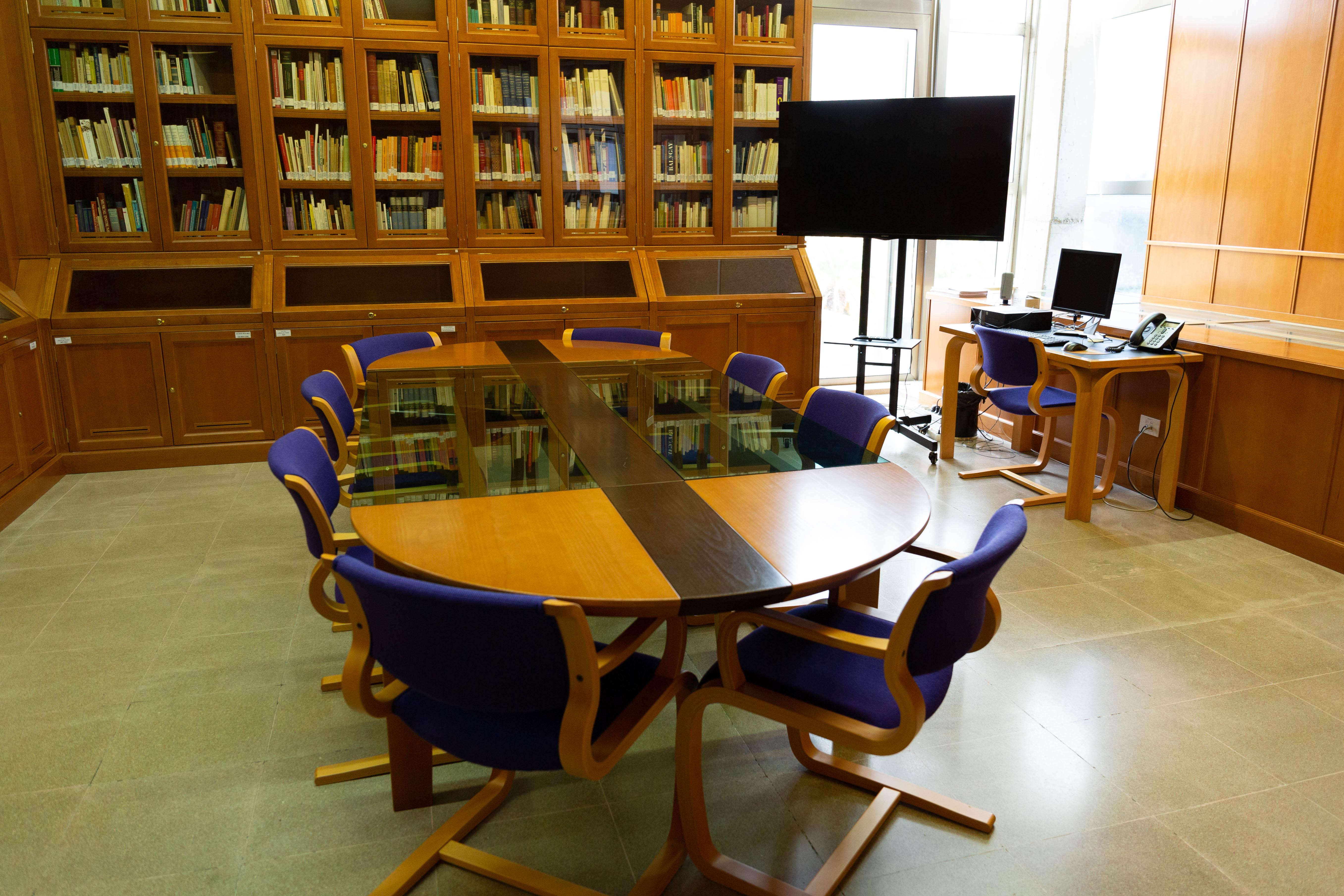 Vista de sala con mesa amplia de reunión con 8 asientos, pantalla gigante, mesa auxiliar con ordenador, pared de estanterías hasta el techo y ventana amplia.