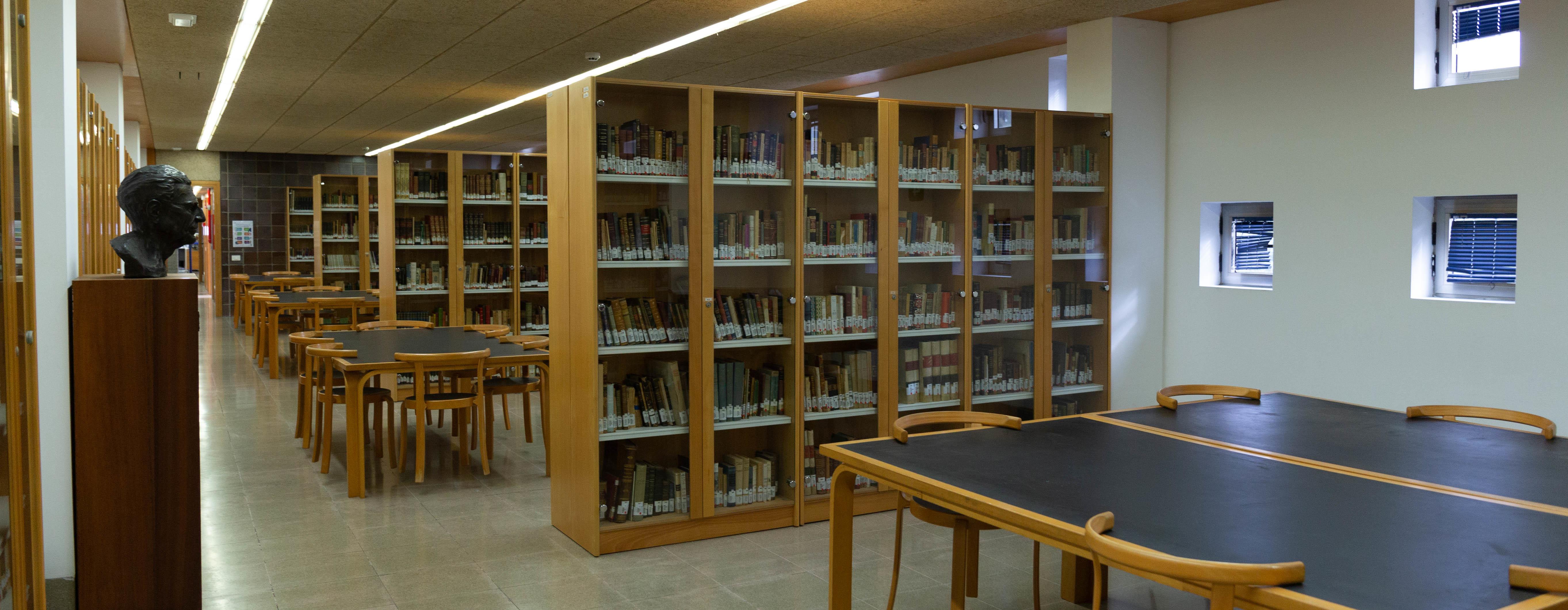 Vista en perspectiva de sala alargada con estanterías de libros que separan espacios dotados de mesas de trabajo y sillas. En un lateral, busto de Saulo Torón.
