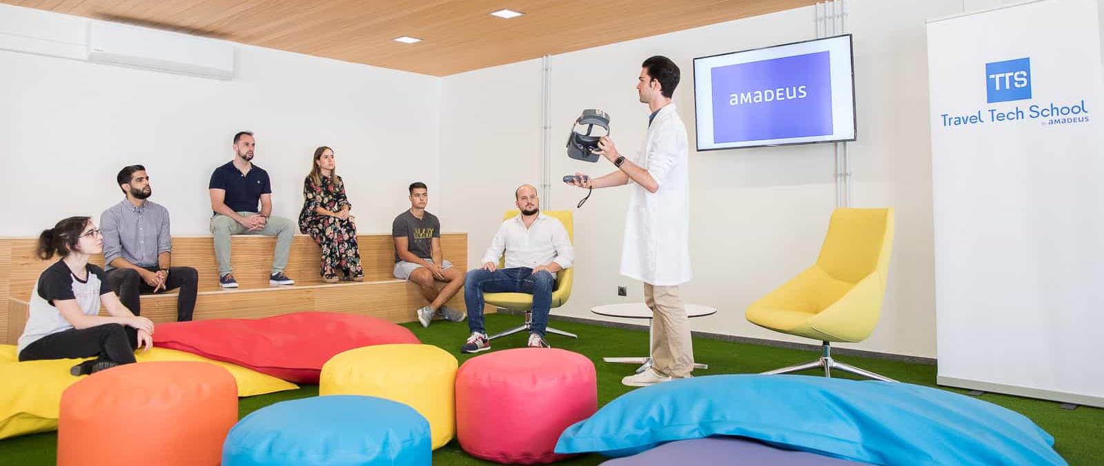 6 personas sentadas en unas pequeñas gradas, un puf y un sillón atieden las explicaciones de una octava, de pie, con bata blanca, que habla mientras muestra unas gafas de 3D en un rincón con césped artificial y cojines gigantes de colores del Travel Tech School de la ULPGC (Edificio Central de la Biblioteca Universitaria)