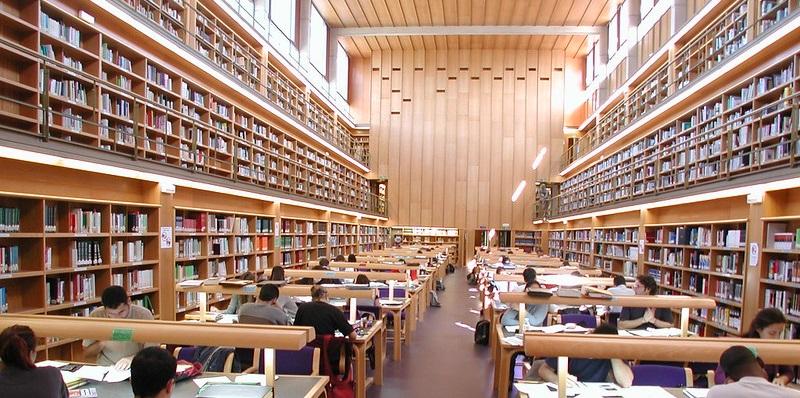 Vista interior de sala de biblioteca alargada, ocupada por dos filas de mesas y con estantes en las paredes laterales, en tres pisos.