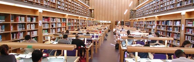 Sala de lecturas con libros, mesas, sillas y personas estudiando